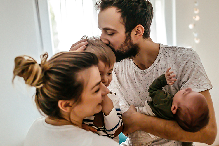 Ein Vater hält ein Baby im Arm und küsst ein Kleinkind auf den Kopf, während eine Mutter lächelnd daneben sitzt. Die Familie wirkt liebevoll und entspannt in einer hellen Wohnumgebung.