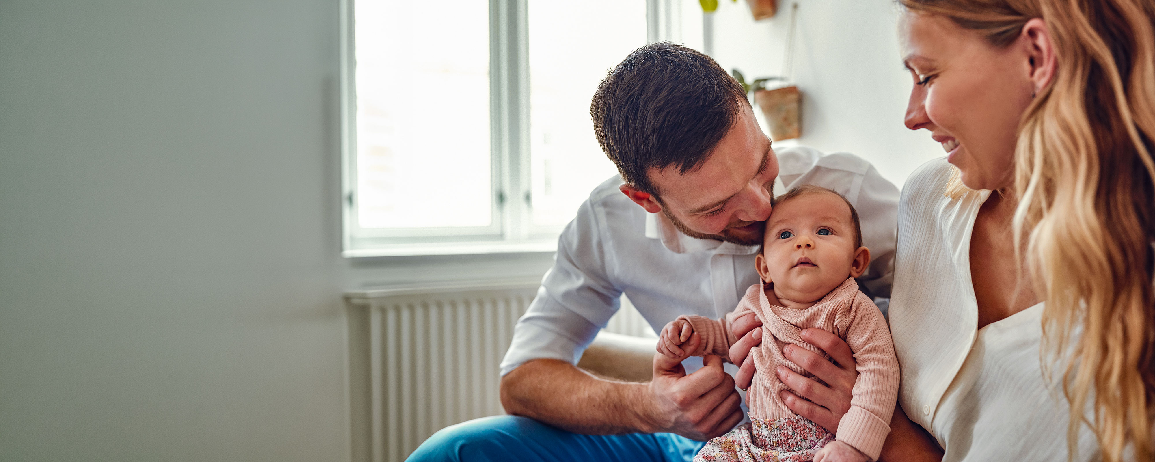 Ein Vater und eine Mutter sitzen zusammen und halten ihr Baby zwischen sich. Der Vater beugt sich vor und küsst das Kind sanft auf den Kopf, während die Mutter lächelnd zuschaut. Die Szene spielt in einem hellen Wohnraum und wirkt liebevoll und geborgen.