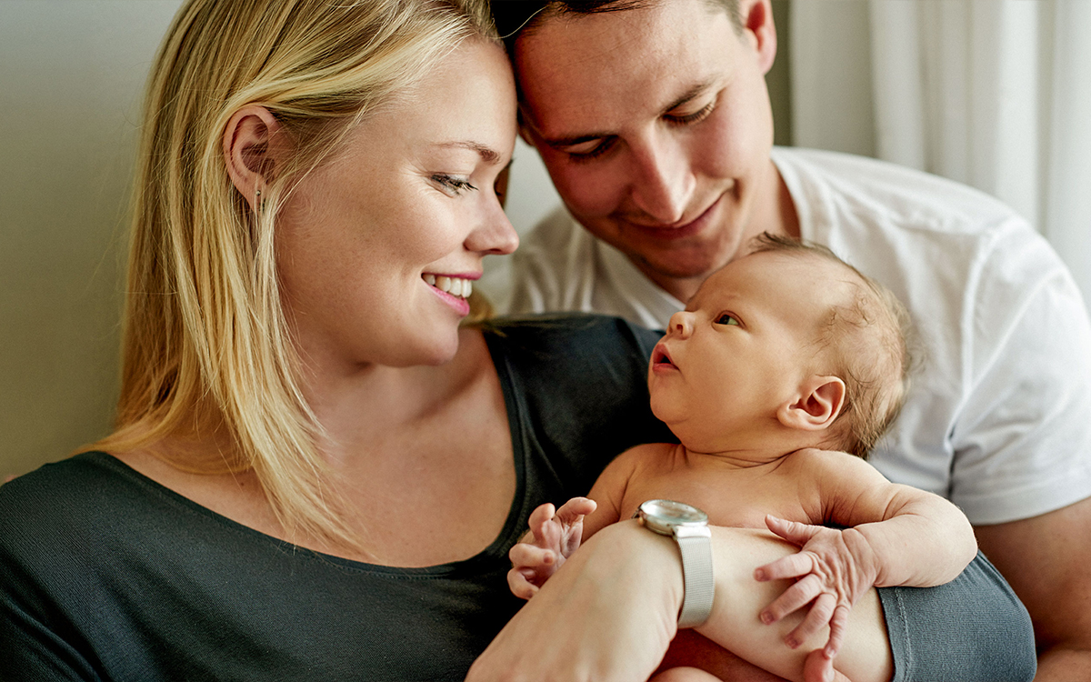 Eine Mutter hält ein Neugeborenes im Arm und lächelt es liebevoll an, während ein Vater sich von hinten dazulehnt und ebenfalls auf das Baby blickt. Die Szene wirkt ruhig, nah und familiär.