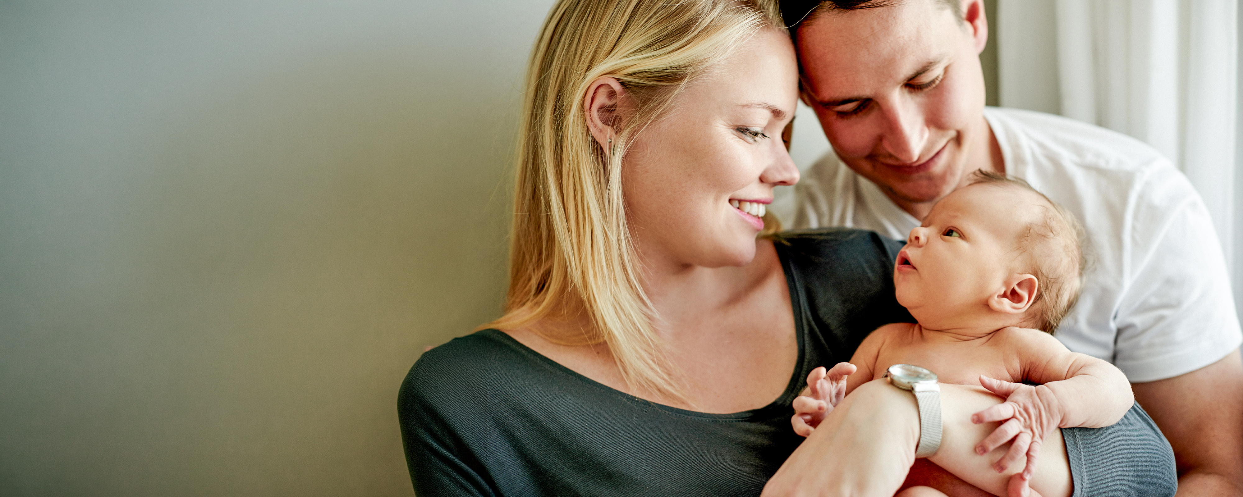 Eine Mutter hält ein Neugeborenes im Arm und lächelt es liebevoll an, während ein Vater sich von hinten dazulehnt und ebenfalls auf das Baby blickt. Die Szene wirkt ruhig, nah und familiär.