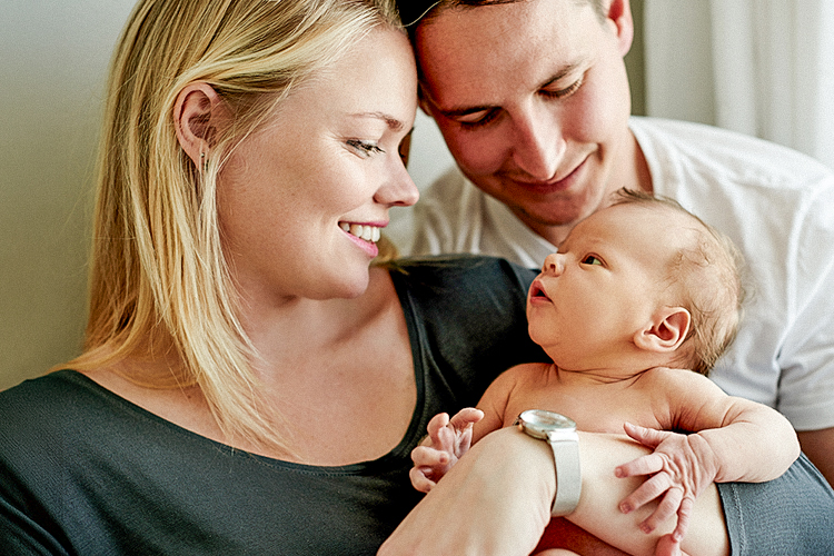Eine Mutter hält ein Neugeborenes im Arm und lächelt es liebevoll an, während ein Vater sich von hinten dazulehnt und ebenfalls auf das Baby blickt. Die Szene wirkt ruhig, nah und familiär.